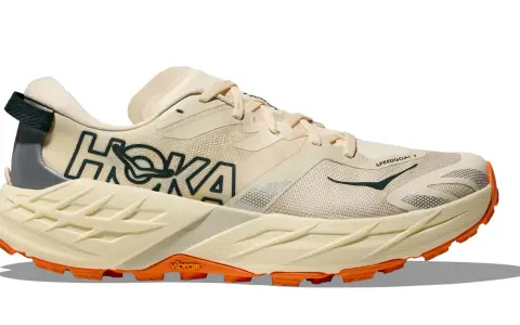 Beiger Hoka Speedgoat 7 Trailrunningschuh mit orangefarbener Vibram-Sohle und dunklem Hoka-Logo.