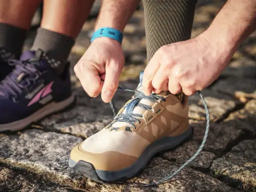 Hände binden Schnürsenkel eines beigefarbenen Schuhs, bereit für den Trail. Daneben ein lila Schuh, der verschiedene wanderschuhe kategorien zeigt.