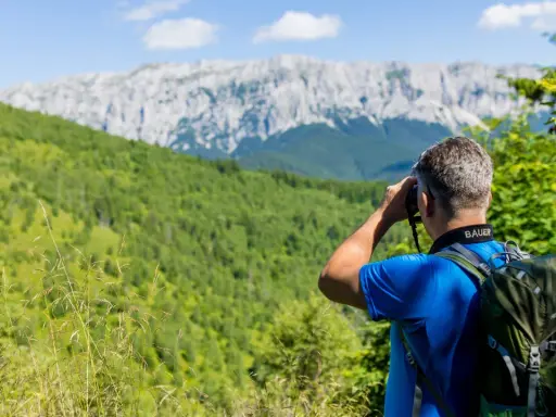 Mann mit Rucksack beim Karpaten Wandern, blickt durch Fernglas auf bewaldete Hänge und felsige Gipfel.