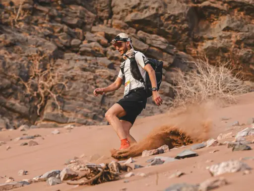 Läufer beim Marathon des Sables wirbelt Sand auf. Ein Mann mit Rucksack und Stirnlampe kämpft sich durch die Wüste.