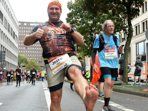 Mann mit schmutzigen Füßen jubelt nach einem Halbmarathon ohne Training. Er hält eine Medaille hoch und gibt Daumen hoch.