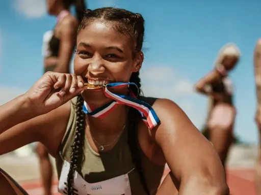Läuferin beißt stolz in ihre Goldmedaille nach einem 10k Lauf. Andere Athletinnen im Hintergrund.