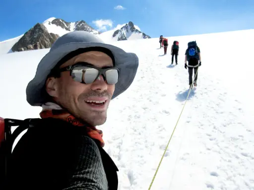 Mann mit Sonnenbrille und Hut, lachend, während er mit einer Gruppe auf einem schneebedeckten Berg wandert. Trail training in den Alpen.