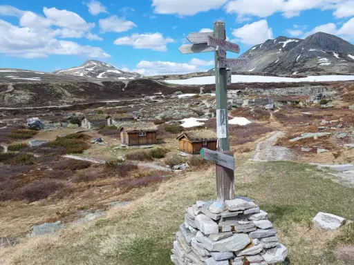 Wanderwegweiser im Rondane Nationalpark, mit grasbewachsenen Hütten und schneebedeckten Bergen im Hintergrund.