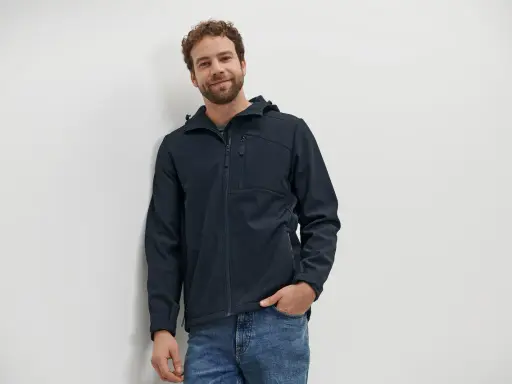 Mann mit lockigem Haar trägt eine dunkelblaue Softshelljacke und Jeans. Die Jacke ist bereit für die Wäsche.