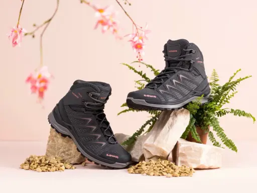 Zwei schwarze Wanderschuhe von LOWA, auf Steinen und Kies arrangiert, mit zarten rosa Blüten und Farn im Hintergrund.