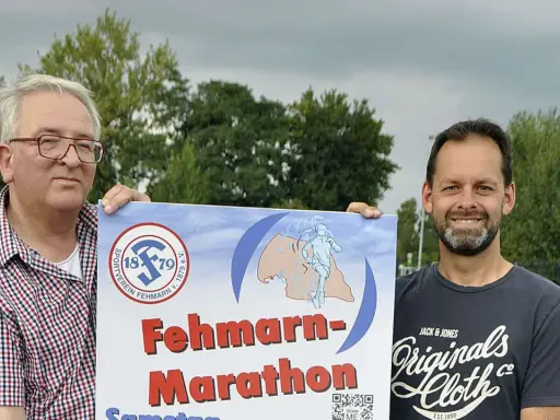 Zwei Männer präsentieren stolz ein Plakat für den Fehmarn Marathon. Das Logo des Sportvereins Fehmarn ist zu sehen.