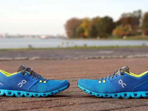 Zwei blaue Laufschuhe, Modell Cloud X4 Damen, stehen auf einem gepflasterten Weg. Im Hintergrund ein See und Bäume.