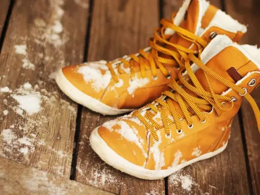 Warme, gefütterte Stiefel mit gelben Schnürsenkeln liegen auf einer Holzplanke, bedeckt mit etwas Schnee. Sie warten darauf, dass die **Schuhe trocknen**.
