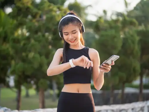 Lächelnde junge Frau mit Kopfhörern und Smartphone, die ihre Smartwatch prüft. Sie trägt atmungsaktive Kleidung für ihr Training im Freien.