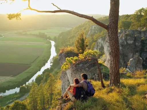 Ein Paar genießt den Sonnenuntergang nach besonderen Wanderungen auf einem Felsvorsprung mit Blick auf ein Tal mit Fluss und Feldern.