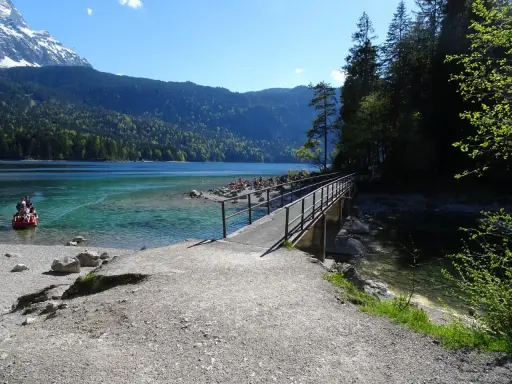 Ein Steg führt über das klare Wasser des Eibsees, ideal für den Eibsee Rundweg. Im Hintergrund Berge und ein rotes Boot.
