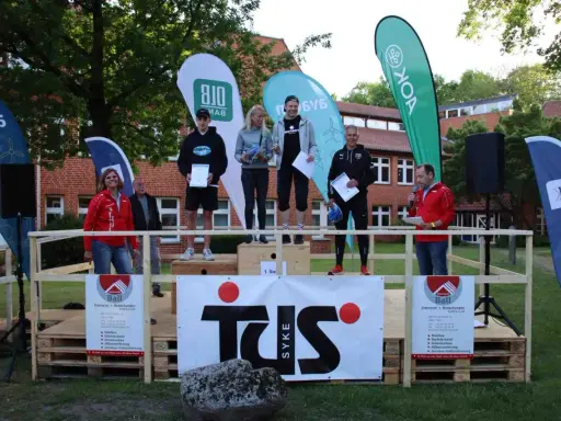 Siegerehrung beim Hachelauf Syke: Läufer auf dem Podium, Moderation und Sponsoren-Banner im Hintergrund.