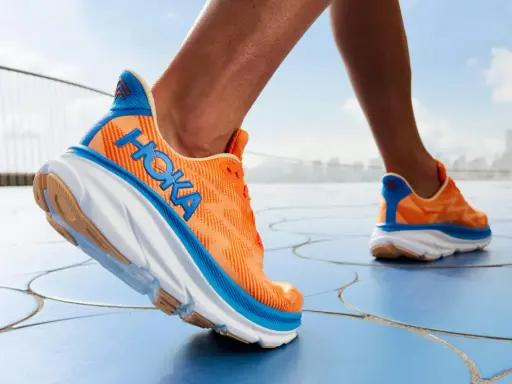 Ein Läufer testet die Hoka Clifton 9 Schuhe in leuchtendem Orange mit blauen Akzenten, die auf einem blauen Boden unterwegs sind.