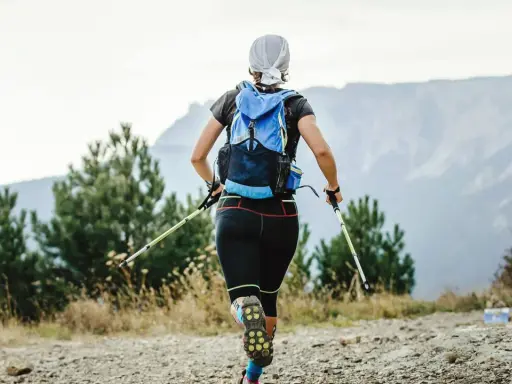 Frauen-Speed-Hiking mit Rucksack und Stöcken auf einem Bergpfad, umgeben von Bäumen und mit Blick auf Berge.