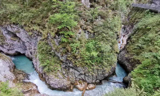 Türkisblaues Wasser rauscht durch eine enge Schlucht mit steilen Felswänden, bewachsen mit üppigem Grün. Eine Holzbrücke führt über die Klammen in Bayern.