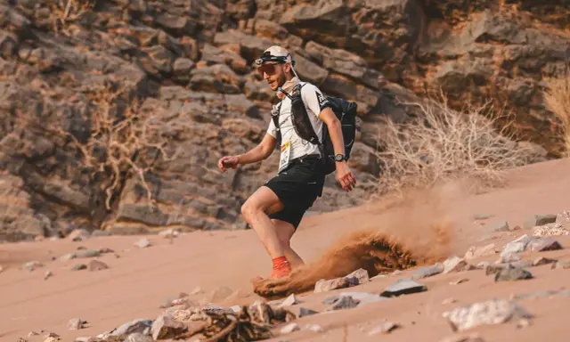 Läufer beim Marathon des Sables wirbelt Sand auf. Ein Mann mit Rucksack und Stirnlampe kämpft sich durch die Wüste.