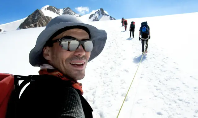 Mann mit Sonnenbrille und Hut, lachend, während er mit einer Gruppe auf einem schneebedeckten Berg wandert. Trail training in den Alpen.
