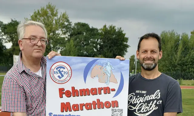 Zwei Männer präsentieren stolz ein Plakat für den Fehmarn Marathon. Das Logo des Sportvereins Fehmarn ist zu sehen.