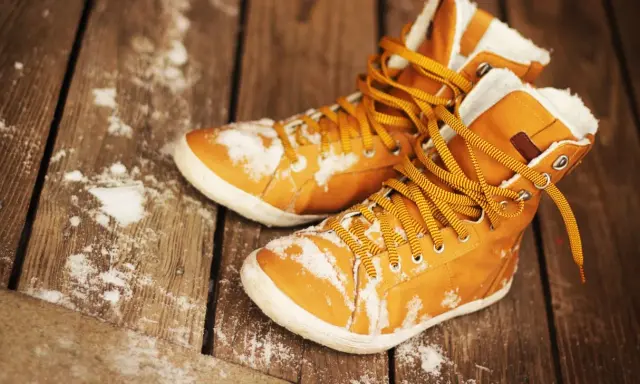 Warme, gefütterte Stiefel mit gelben Schnürsenkeln liegen auf einer Holzplanke, bedeckt mit etwas Schnee. Sie warten darauf, dass die **Schuhe trocknen**.