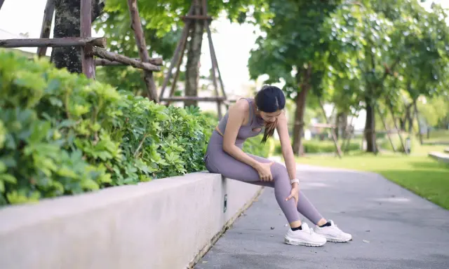 Frau sitzt im Park, hält ihr Bein und scheint Schmerzen zu haben. Mögliche Ursache: schwere Beine beim Laufen.
