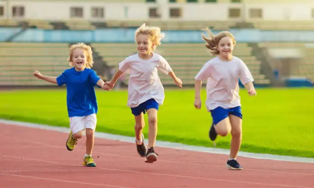 Drei lachende Kinder beim Lauftraining auf einer roten Laufbahn. Sie rennen nebeneinander, die Arme ausgestreckt.