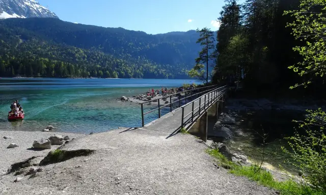 Ein Steg führt über das klare Wasser des Eibsees, ideal für den Eibsee Rundweg. Im Hintergrund Berge und ein rotes Boot.