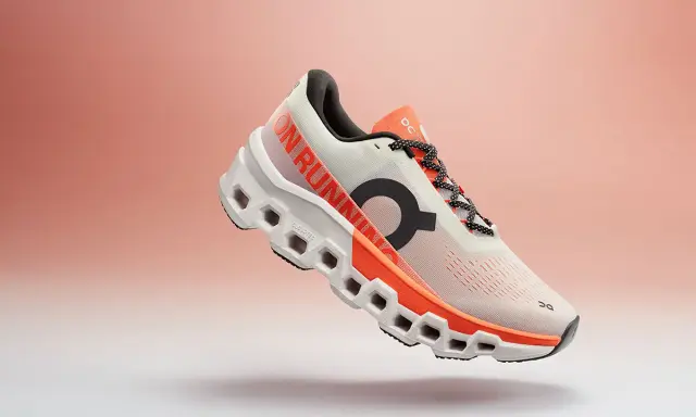 Ein weißer Laufschuh mit orangefarbenen Akzenten, der Cloudmonster 2 Herren, schwebt vor einem sanften Farbverlauf.