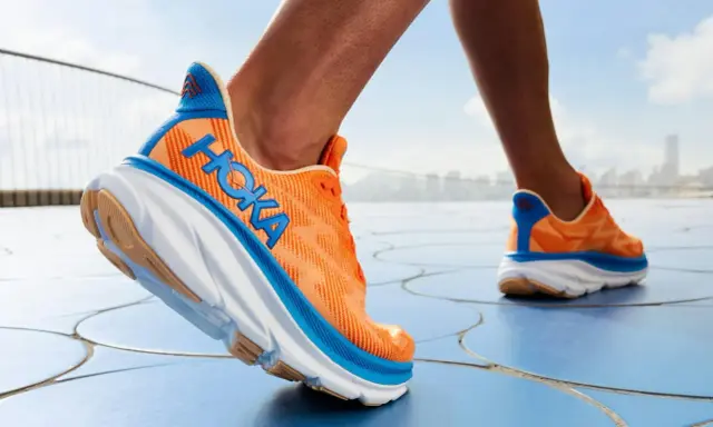 Ein Läufer testet die Hoka Clifton 9 Schuhe in leuchtendem Orange mit blauen Akzenten, die auf einem blauen Boden unterwegs sind.