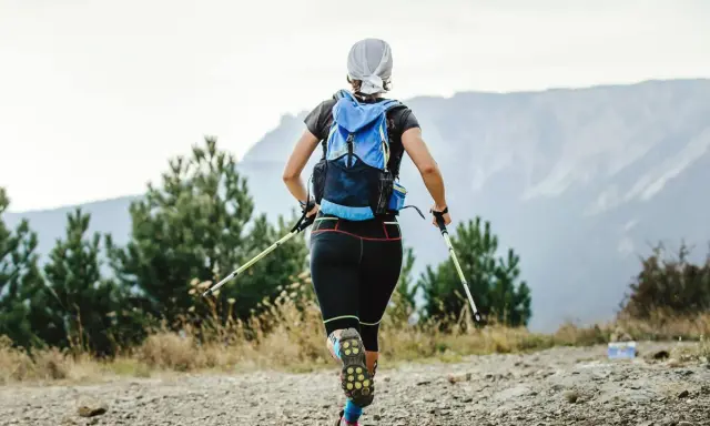 Frauen-Speed-Hiking mit Rucksack und Stöcken auf einem Bergpfad, umgeben von Bäumen und mit Blick auf Berge.