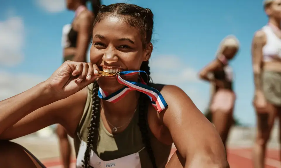 Läuferin beißt stolz in ihre Goldmedaille nach einem 10k Lauf. Andere Athletinnen im Hintergrund.