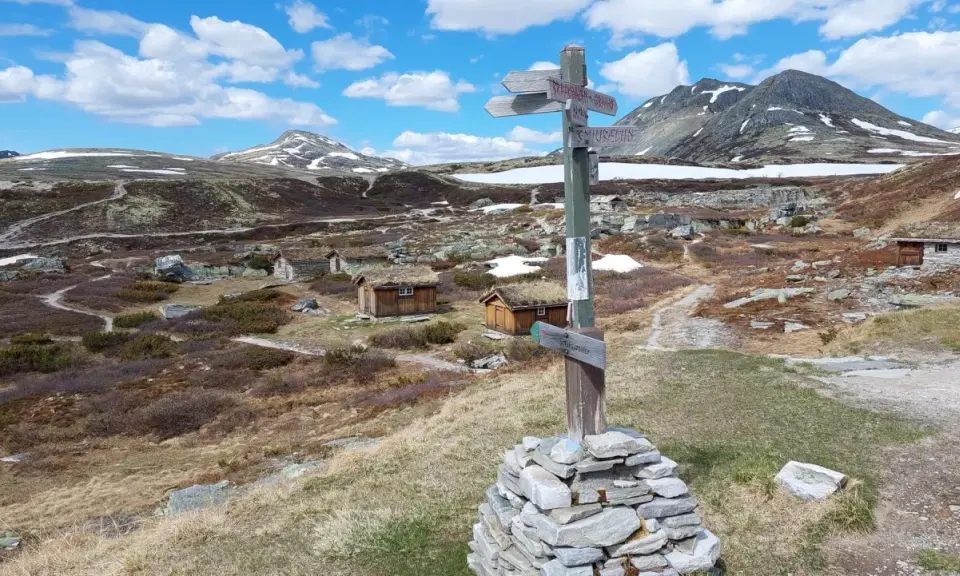 Wanderwegweiser im Rondane Nationalpark, mit grasbewachsenen Hütten und schneebedeckten Bergen im Hintergrund.