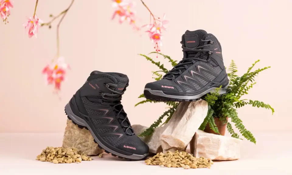 Zwei schwarze Wanderschuhe von LOWA, auf Steinen und Kies arrangiert, mit zarten rosa Blüten und Farn im Hintergrund.