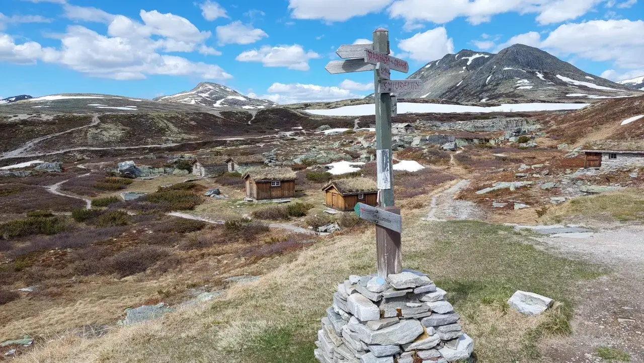 Wanderwegweiser im Rondane Nationalpark, mit grasbewachsenen Hütten und schneebedeckten Bergen im Hintergrund.