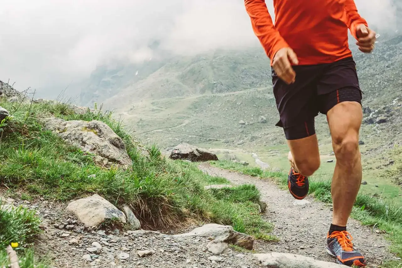 Ein Läufer im orangen Shirt und schwarzen Shorts absolviert sein trailrunning training auf einem steinigen Bergpfad.