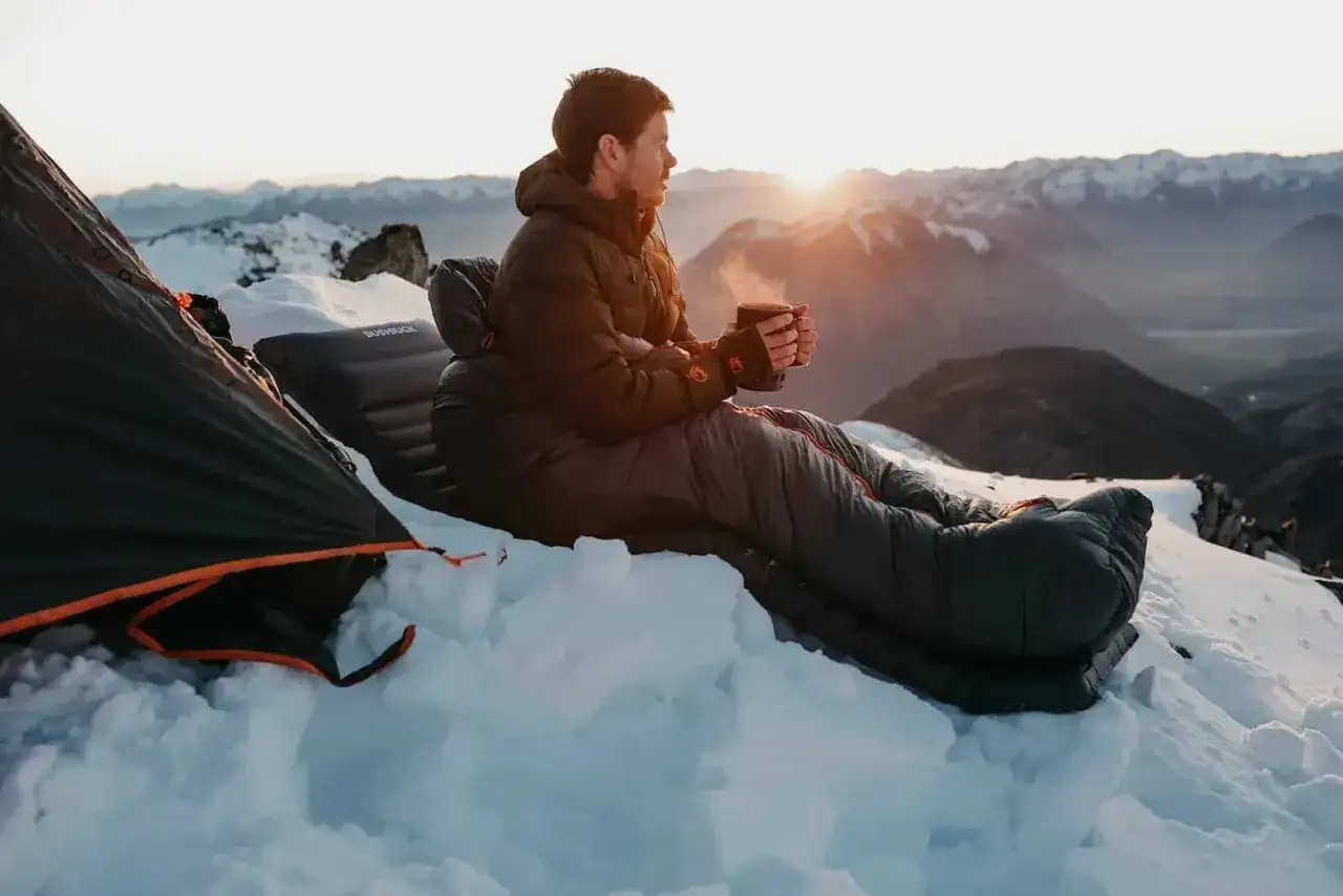Mann genie&szlig;t hei&szlig;en Tee im Schlafsack auf verschneiter Bergspitze. Die 600 cuin F&uuml;llung h&auml;lt ihn warm, w&auml;hrend die Sonne &uuml;ber den Gipfeln aufgeht.