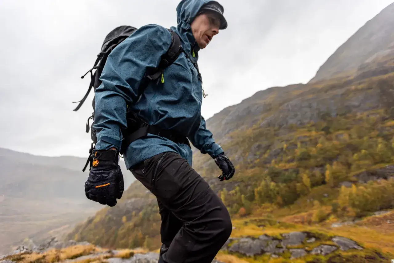 Wanderer in Regenjacke und Handschuhen, unterwegs in bergiger Landschaft. Die Ausr&uuml;stung ist PFC frei und sch&uuml;tzt vor N&auml;sse.