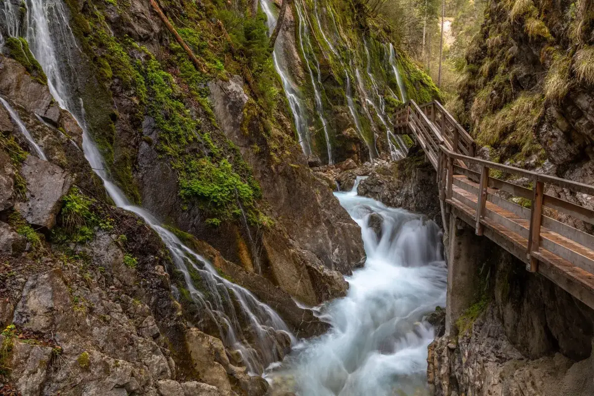 Ein rauschender Bach flie&szlig;t durch eine Schlucht mit vielen kleinen Wasserf&auml;llen, die von moosbewachsenen Felsen herabst&uuml;rzen. Ein Holzsteg f&uuml;hrt entlang der Klammen in Bayern.