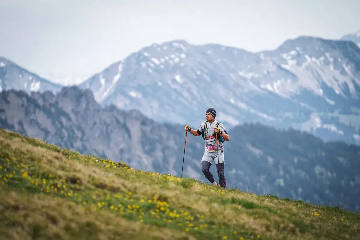Ein Mann, ein mountainman nesselwang, wandert mit Stöcken auf einem blumenübersäten Hang. Im Hintergrund erheben sich majestätische, schneebedeckte Berge.