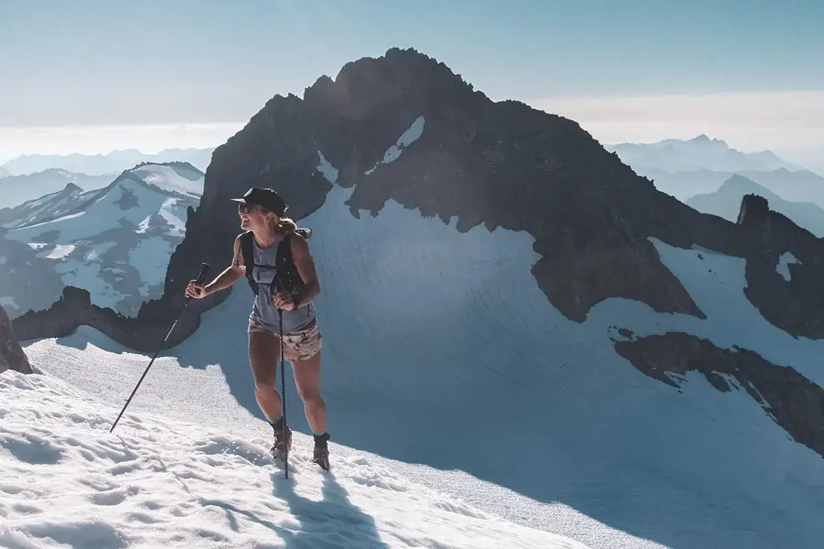 Frau beim Skyrunning auf schneebedecktem Berggipfel, mit St&ouml;cken und Rucksack, im Hintergrund weitere Berge.