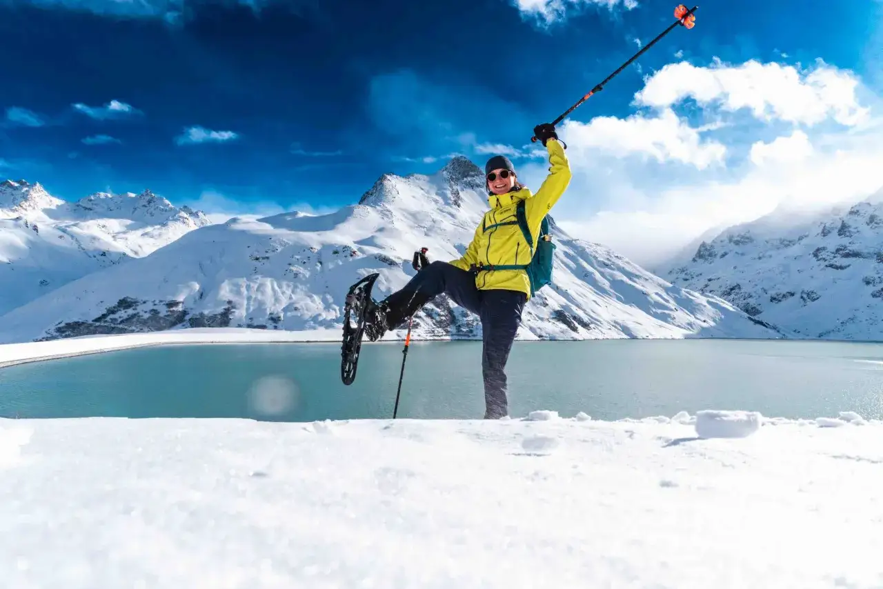 Glückliche Person mit Schneeschuhwandern Schuhen und Stöcken feiert auf verschneiter Bergspitze vor blauem See und schneebedeckten Gipfeln.