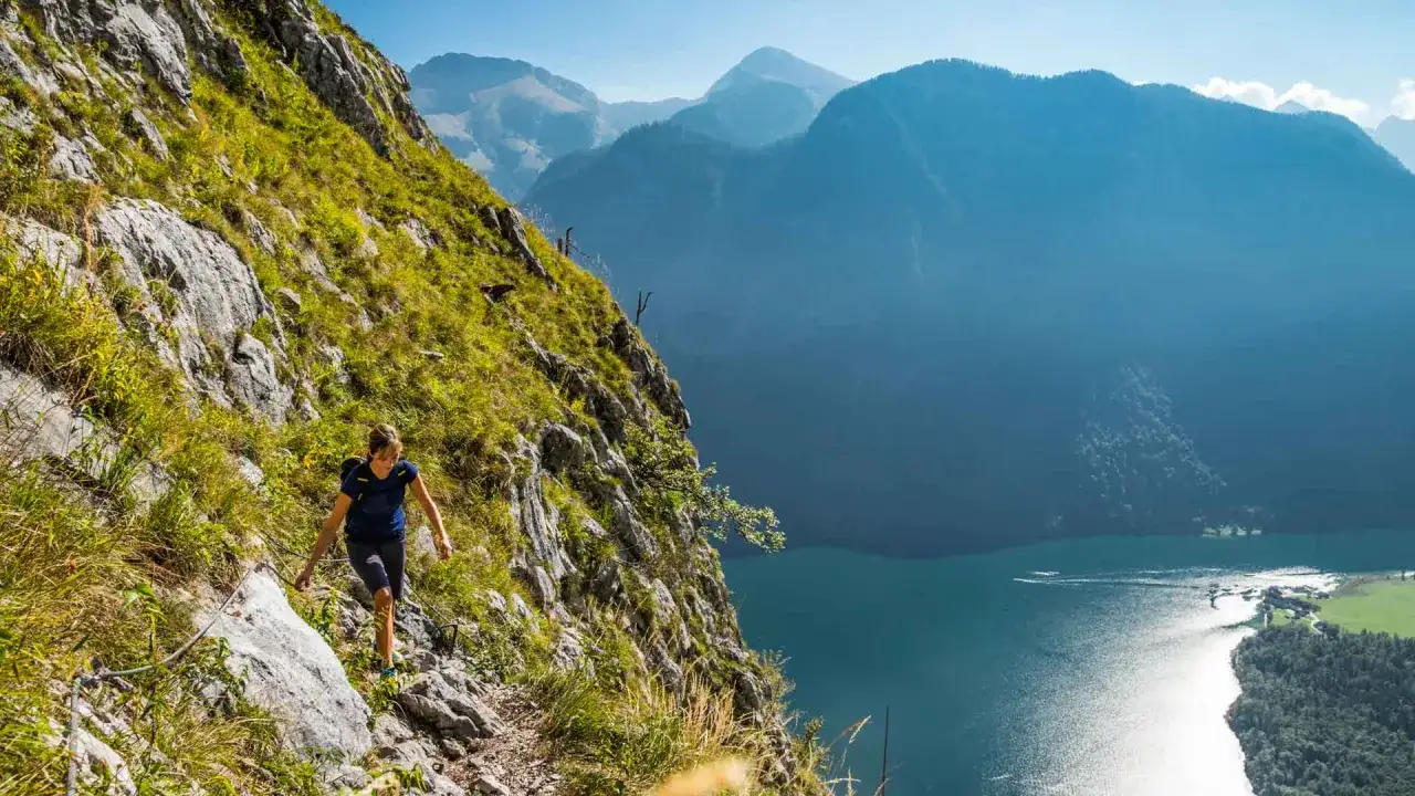 Wanderin auf einem steilen Pfad mit Seilsicherung, Blick auf den Königssee. Die Königssee Umrundung bietet atemberaubende Ausblicke auf Berge und See.
