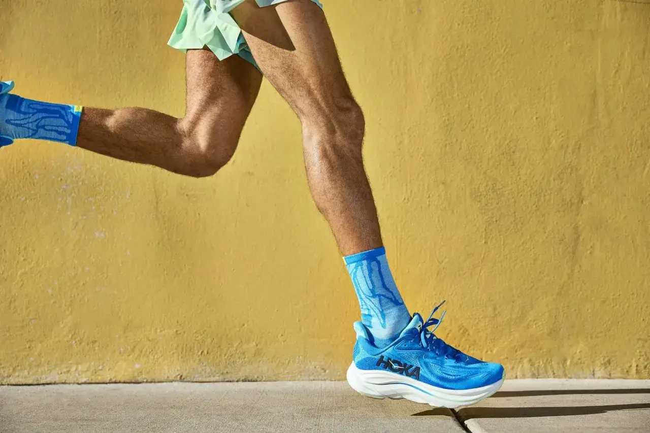 Läufer mit blauen Hoka Schuhen und Socken vor gelber Wand. Positive Hoka Schuhe Erfahrungen sind garantiert.
