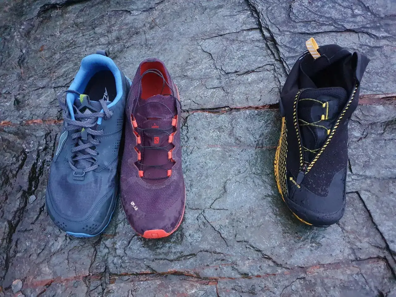 Drei Wanderschuhe liegen auf nassem Fels. Der linke ist blau, der mittlere lila. Der rechte schwarze Schuh hat eine gelbe Schnürung und Gore-Tex. Vibram Sohle Nachteile sind hier nicht ersichtlich.