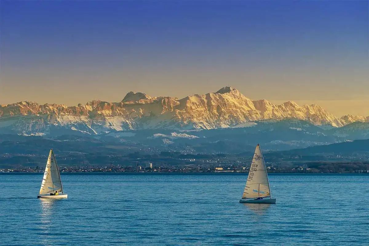 Zwei Segelboote auf dem Bodensee, im Hintergrund schneebedeckte Berge. Eine perfekte Kulisse für eine Wanderung am See.