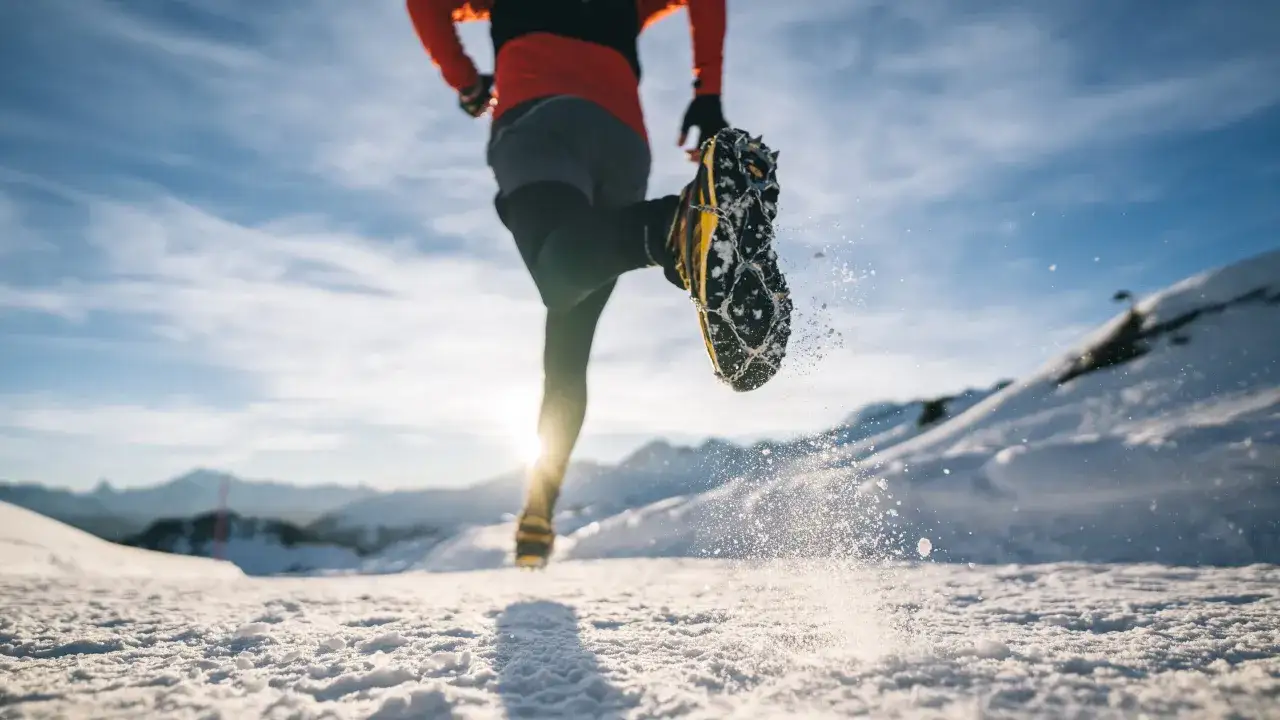 Läufer mit Spikeschuhen im Schnee, bereit für Trailrunning Training. Die Sonne scheint hinter den Bergen.