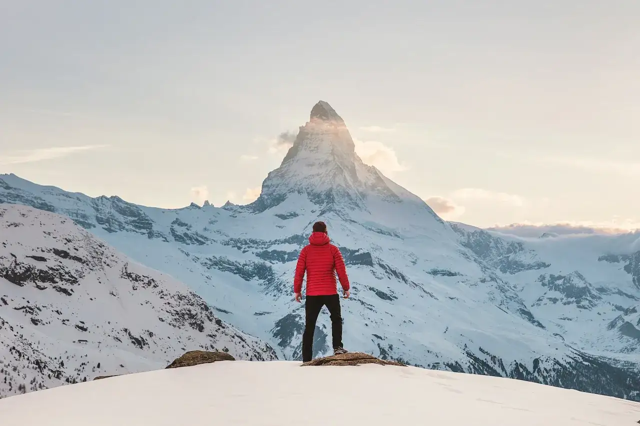 Mann in roter Winterjacke und Hose genie&szlig;t den Ausblick auf schneebedeckte Berge. Perfekte Kleidung f&uuml;rs Wandern im Winter.