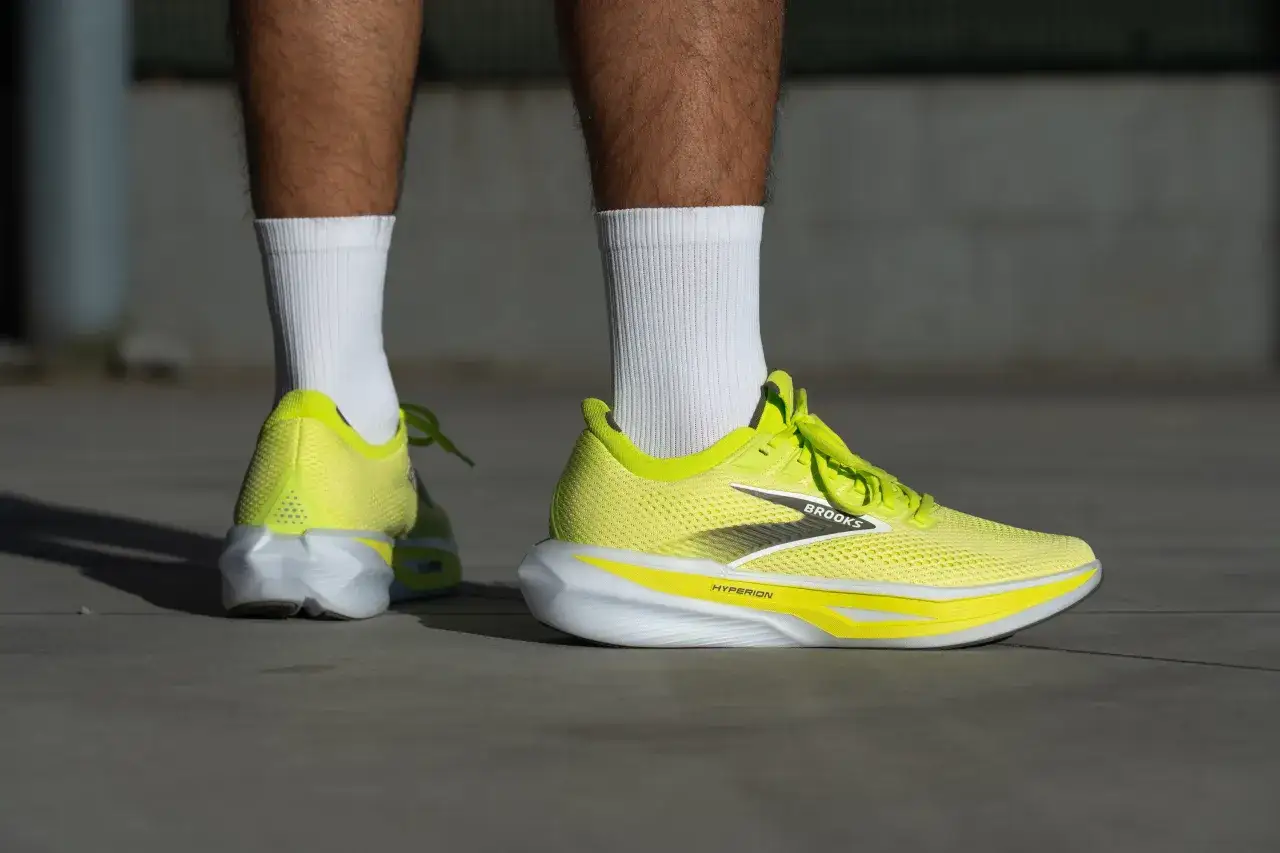 Leuchtend gelbe Brooks Hyperion 3 Laufschuhe mit wei&szlig;en Socken auf grauem Untergrund.