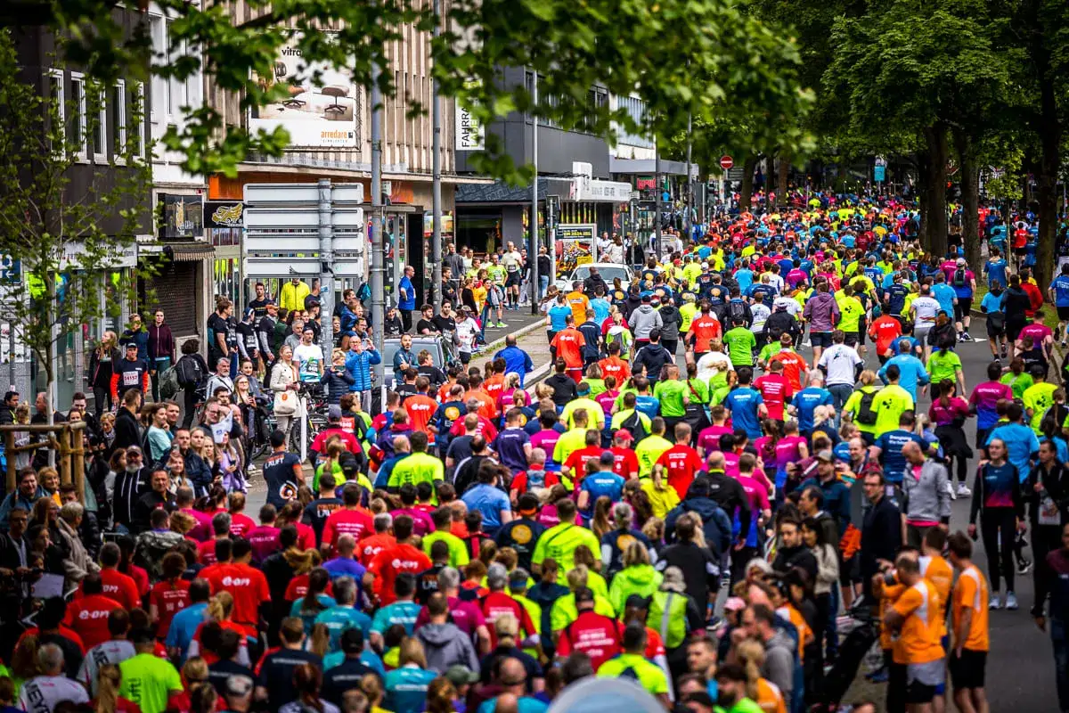 Gro&szlig;e Menschenmenge beim Firmenlauf Essen, viele L&auml;ufer in bunten Trikots auf einer Stra&szlig;e, umgeben von B&auml;umen und Geb&auml;uden.