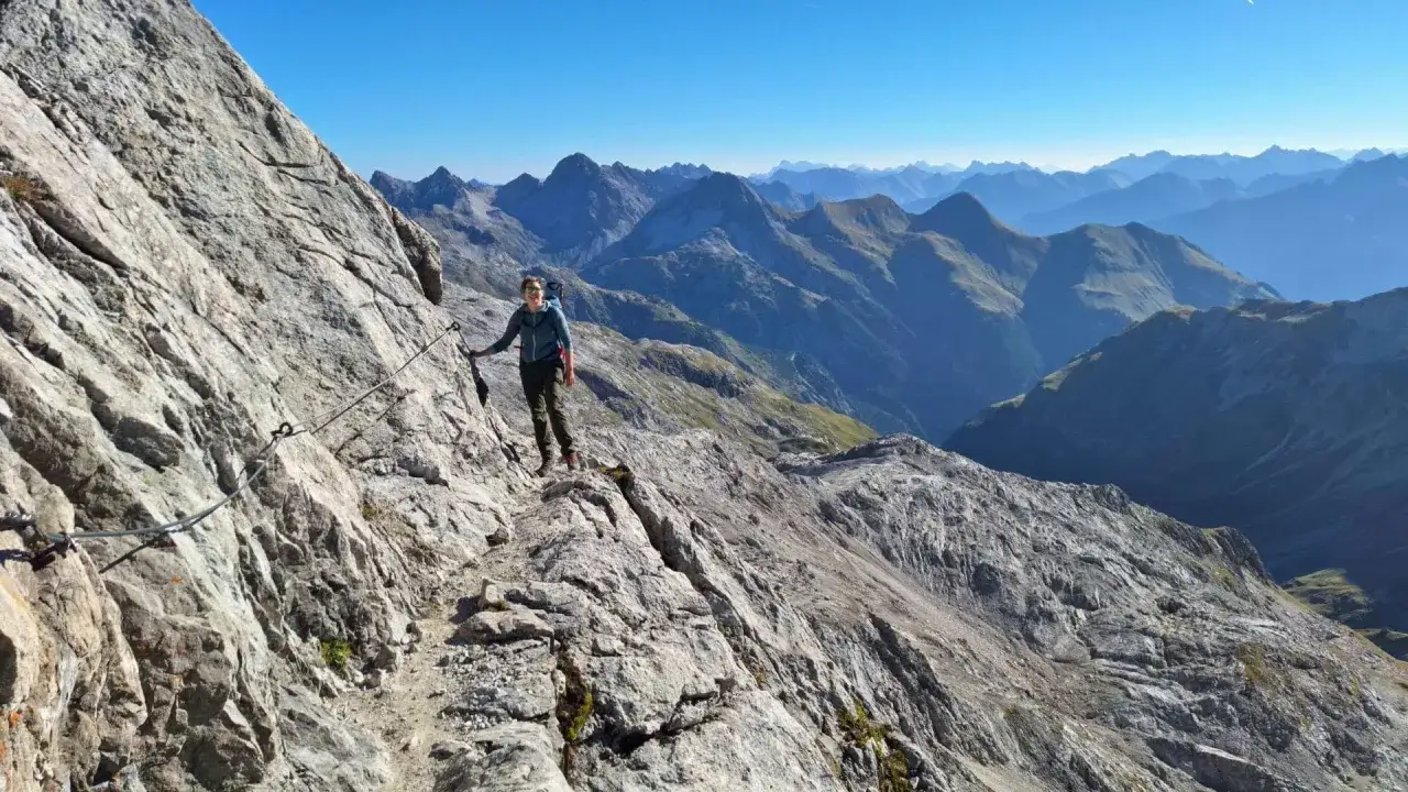 Frau auf dem Heilbronner Weg wandern, gesichert an einem Stahlseil, mit atemberaubendem Bergpanorama im Hintergrund.